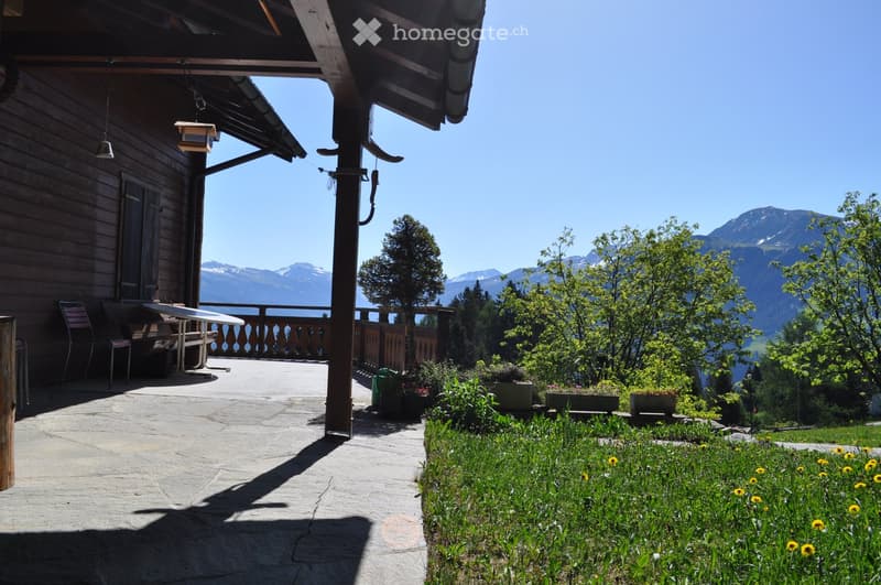 A wooden deck area with a table and chairs, overlooking a scenic mountain view with lush greenery and flowers.