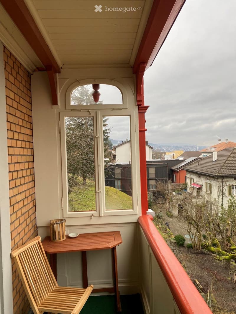 Balcony with wooden table, chair, candle holder, and dish, overlooking a residential area with trees and houses.