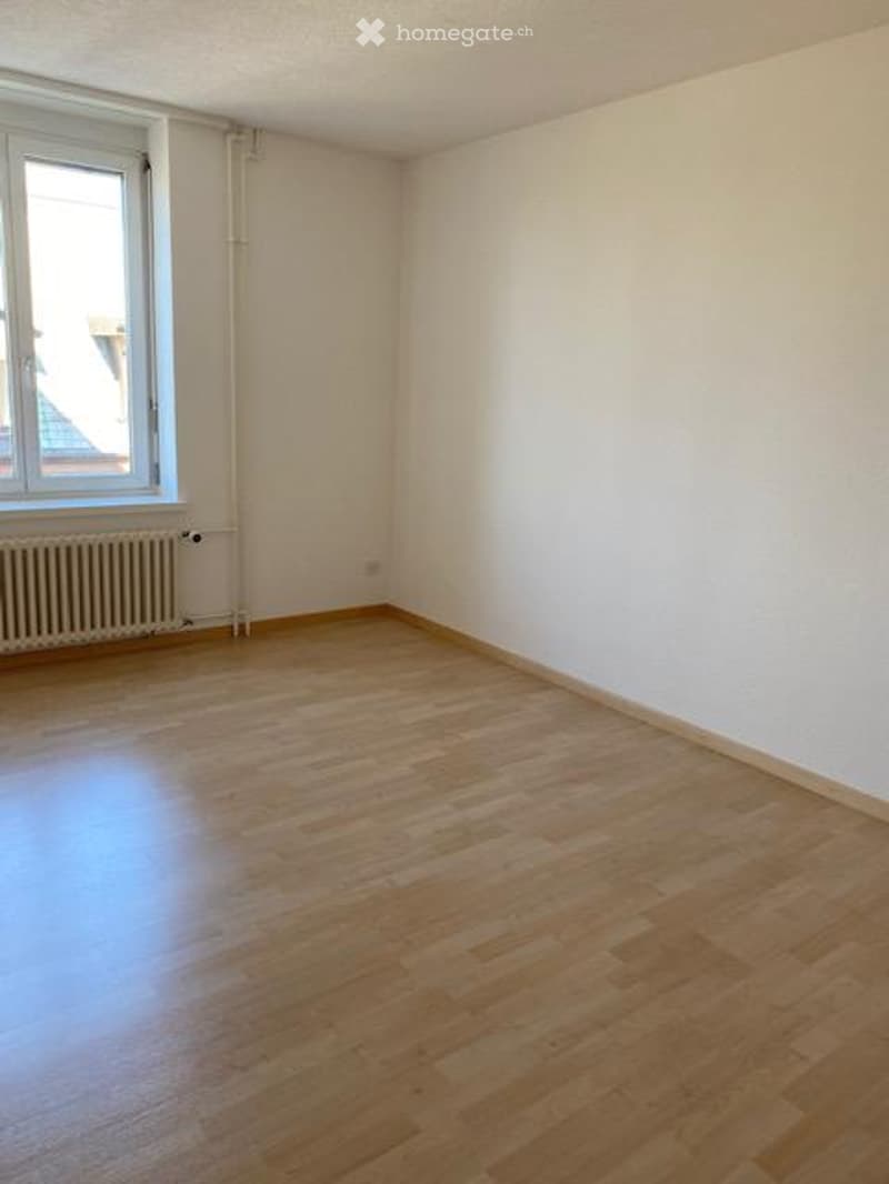 Empty room with wooden flooring, white walls, and a single window with radiator below.