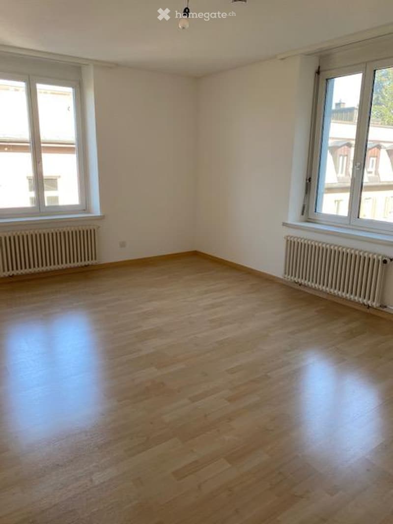 Empty room with wooden flooring, white walls, and large windows with radiators underneath.