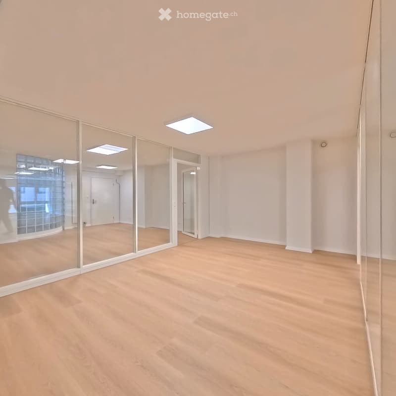 Empty room with light wooden flooring, white walls, and large glass partitions. No furniture or equipment present.