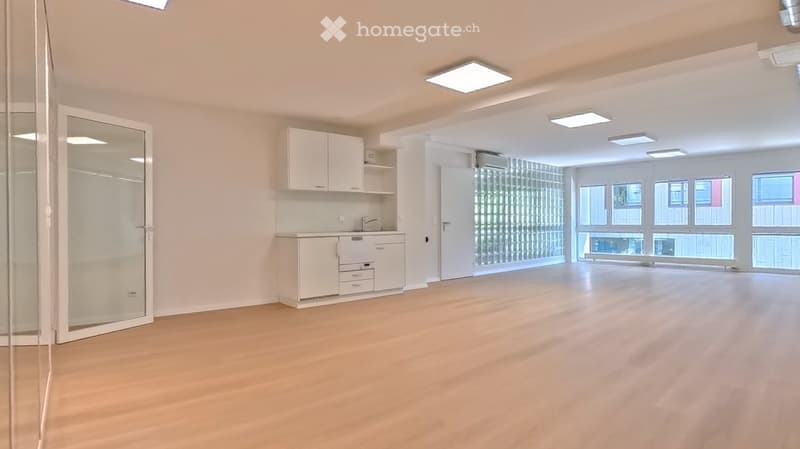 Empty room with light wooden flooring, white walls, and a small kitchenette area with cabinets and a sink. Large windows provide natural light.