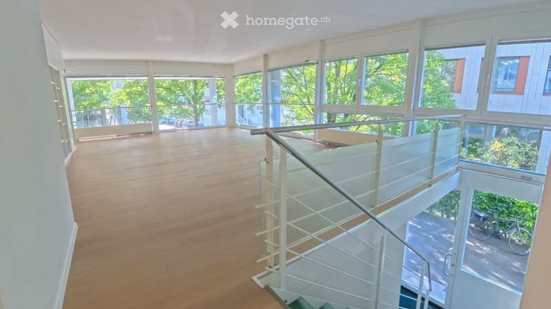 Open, empty room with wooden flooring, large windows, and a modern glass staircase leading to an upper level.