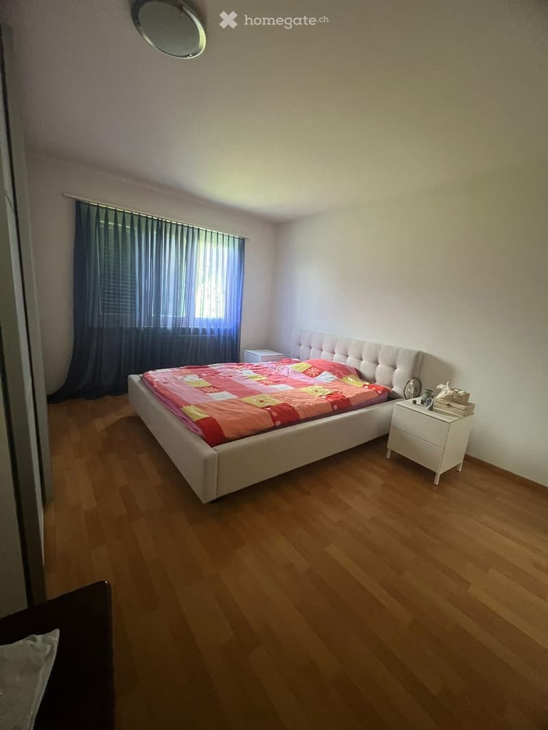 Bedroom with wooden flooring, a large bed with a colorful quilt, a nightstand, and a window with dark curtains.