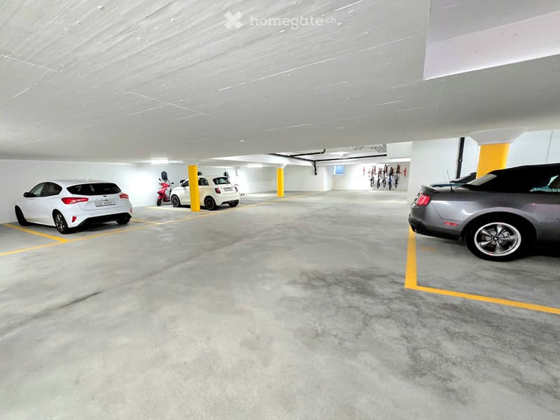 Underground parking area with several parked cars, including a white hatchback, a grey SUV, and a scooter. The space features concrete floors, yellow parking lines, and yellow support pillars.