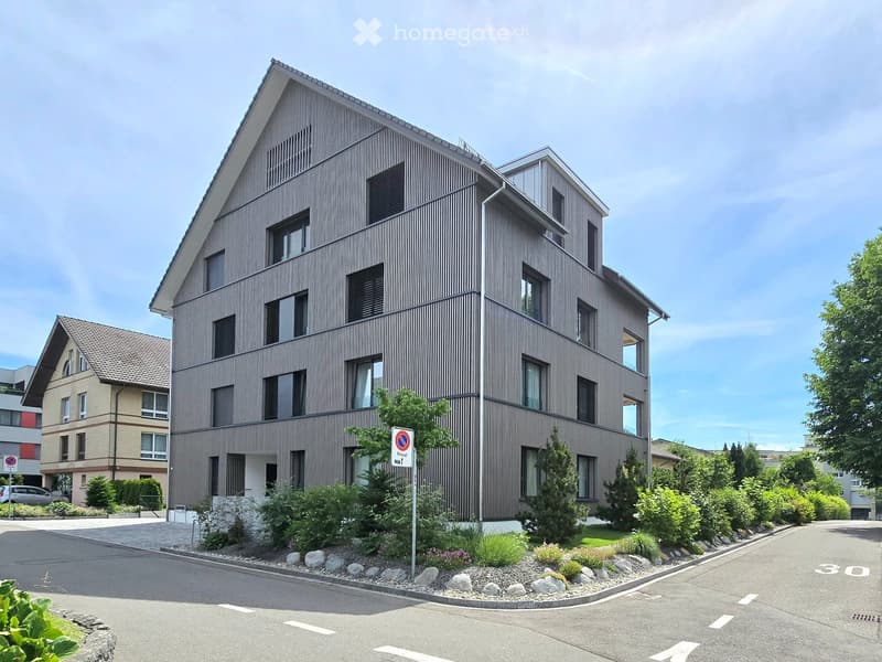 Modern multi-story residential building with a contemporary facade, large windows, and a landscaped entrance area with shrubs and rocks.
