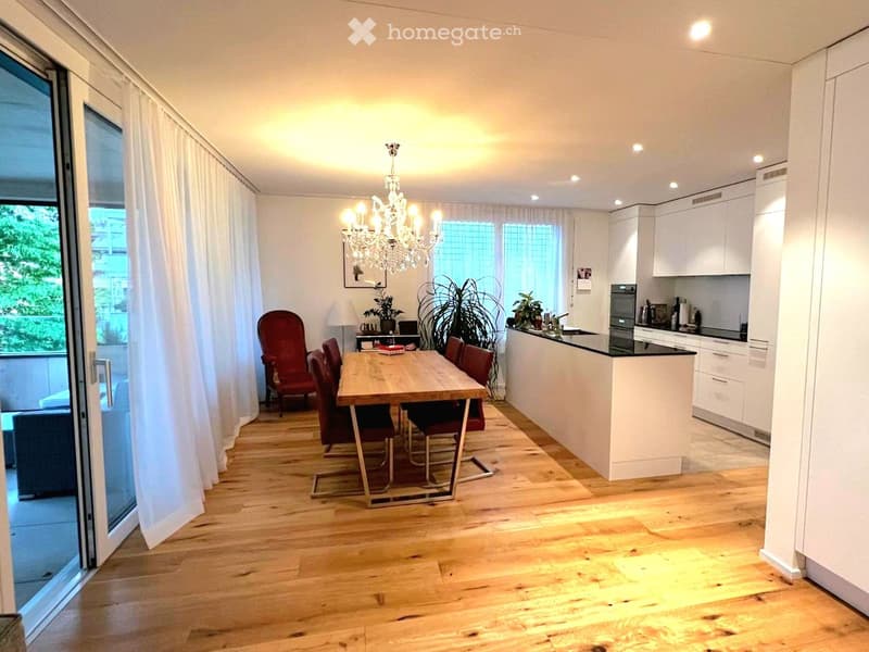Open-plan living and dining area with wooden flooring, large windows, a dining table with chairs, a kitchen island, and a chandelier.