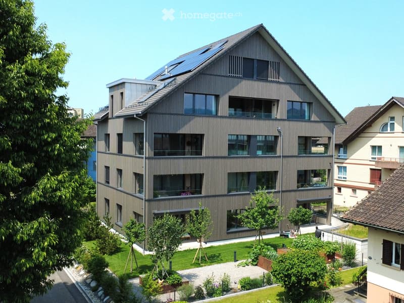 Modern multi-story residential building with large windows, balconies, and solar panels on the roof, surrounded by a well-maintained garden and trees.