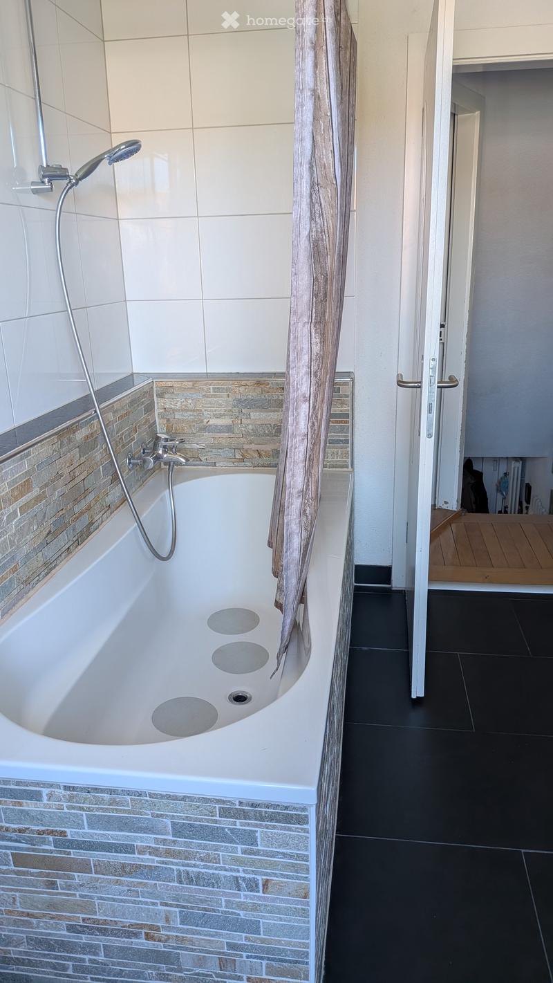 Modern bathroom with a white bathtub, tiled walls, and a showerhead attached to the wall. The floor is dark tiled, and a wooden door is visible in the background.