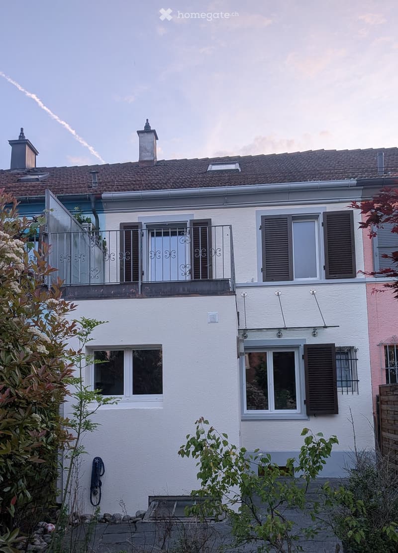 Two-story house with white and pink exterior, multiple windows with shutters, a balcony with metal railing, and a small garden area with plants and a hose reel.