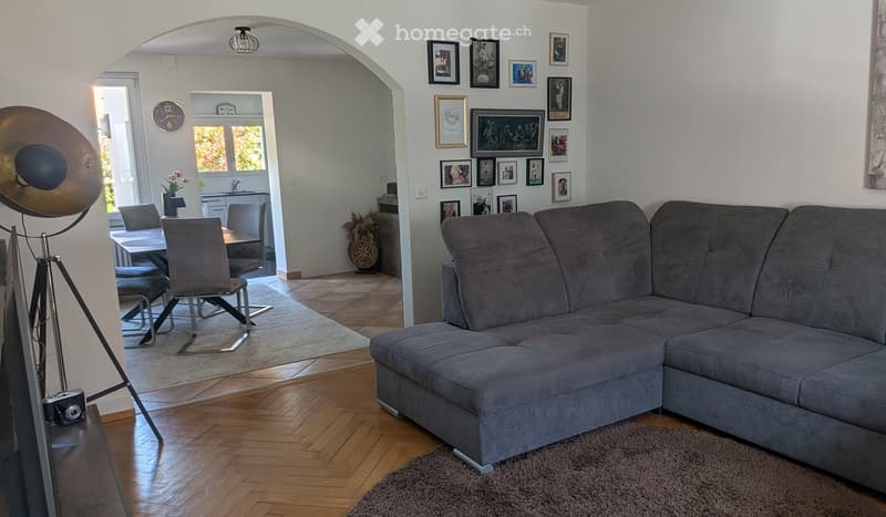 Modern living room with a grey sectional sofa, wooden floor, dining area with table and chairs, and wall art.