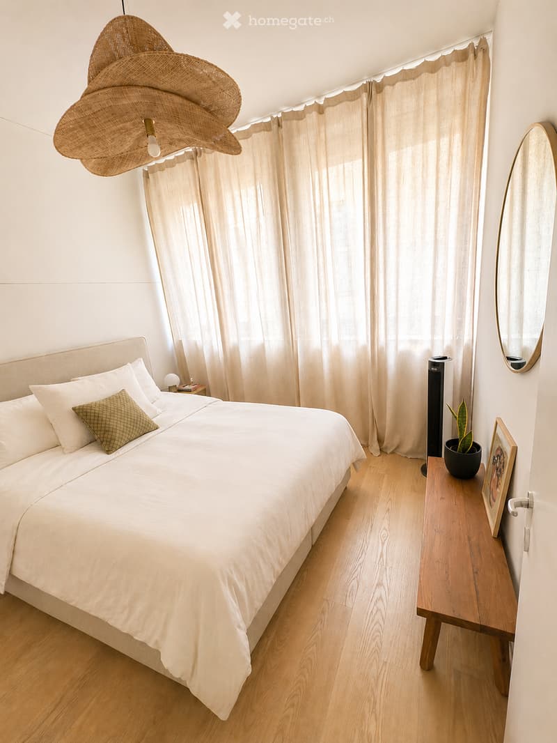 A modern, well-lit bedroom with a large bed, white bedding, a nightstand, a mirror, a small table with a potted plant, and a decorative picture frame. The room features light wooden flooring and large windows with sheer curtains.