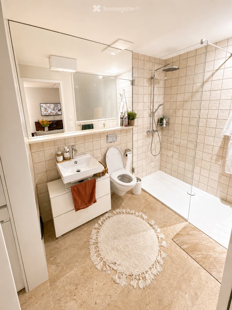 Modern bathroom with beige tiles, a white sink, toilet, shower, and a large mirror. Features a fluffy white rug, a towel, and a small plant on the shelf.
