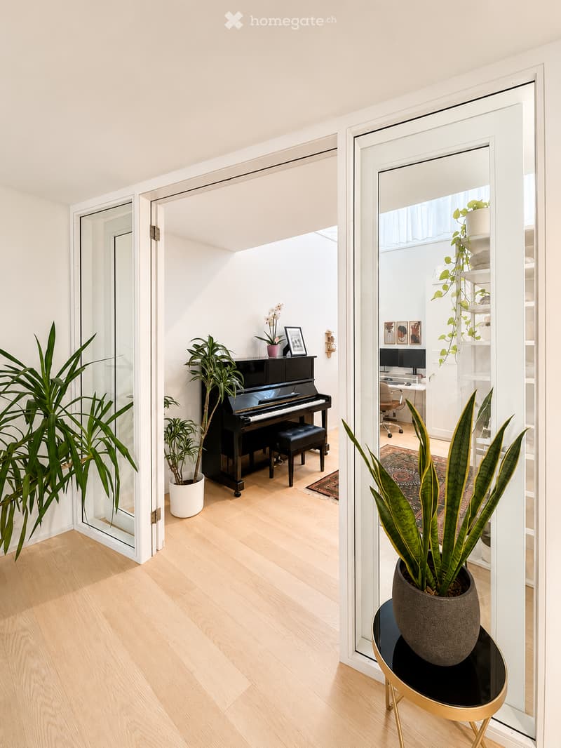 A modern, well-lit room with light wooden flooring, featuring a grand piano, a small round table with a potted plant, and a workspace area with a desk, chair, and computer screen.