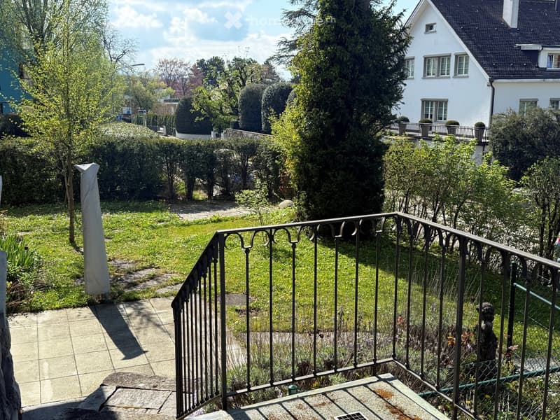 Balcony overlooking a well-maintained garden with green grass, trees, shrubs, and a pathway leading to a house in the background.