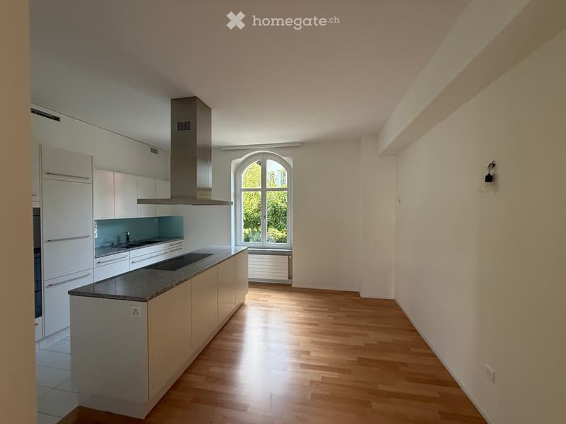 Modern kitchen with white cabinets, a central island, and a large window letting in natural light. The floor is light wooden, and the countertops are dark grey.