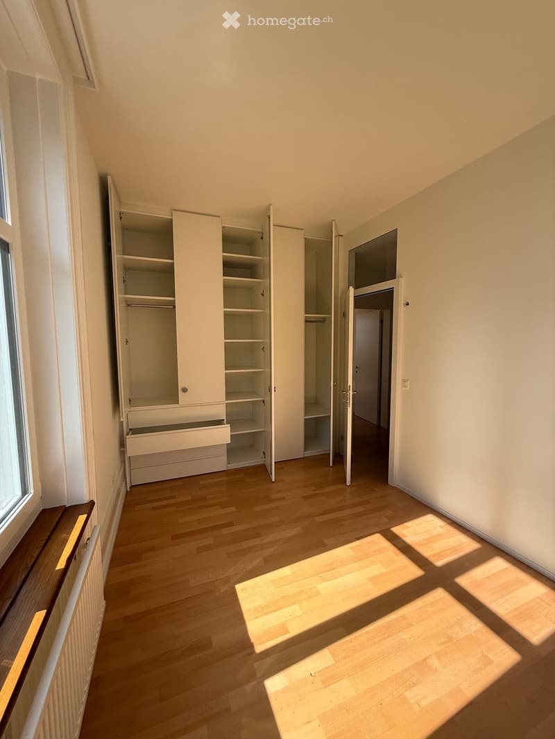 Empty room with wooden flooring, white walls, built-in closet with shelves and drawers, and a window letting in natural light.