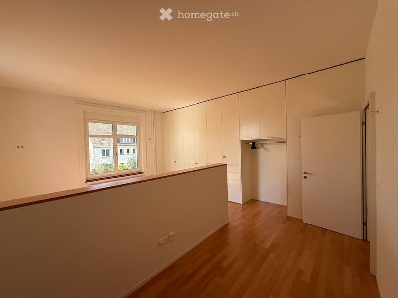 Empty room with light wooden flooring, white walls, built-in wardrobes, and a window offering a view of neighboring buildings.