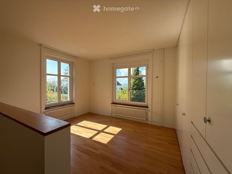 Empty room with wooden flooring, large windows, radiators, and a wooden railing visible on the left side.
