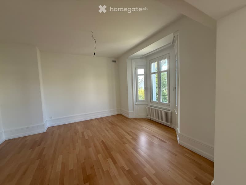 Empty room with wooden flooring, white walls, and large windows with white frames. A radiator is visible under one window.