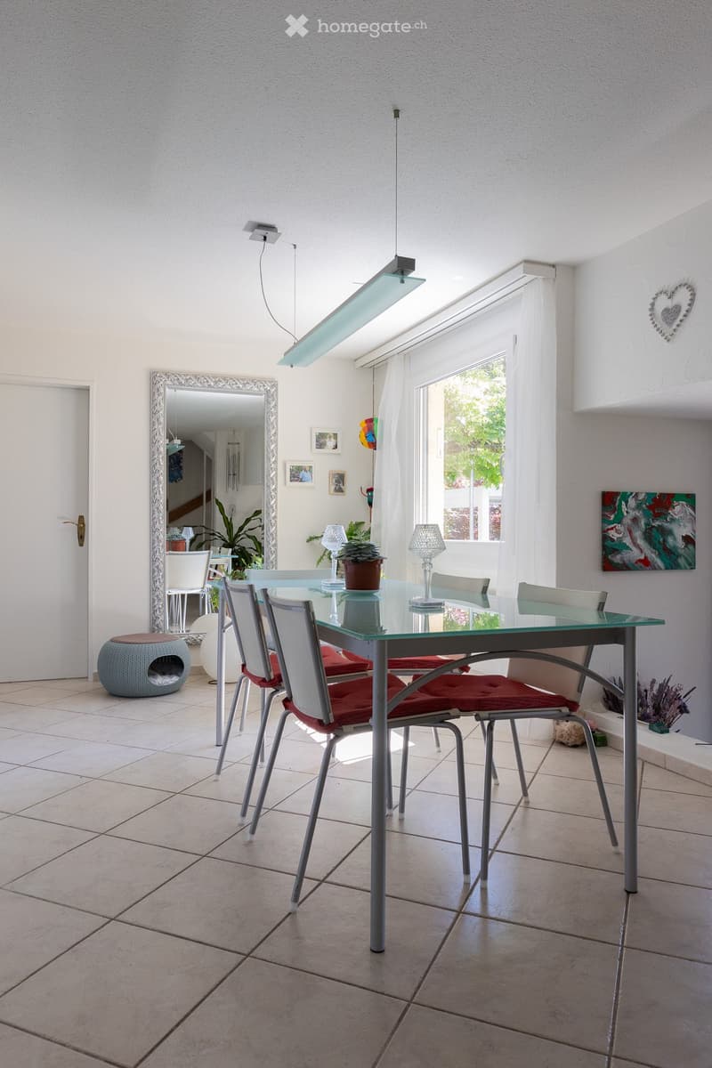 Bright dining area with a glass table, red cushioned chairs, a large mirror, a decorative stool, and potted plants.