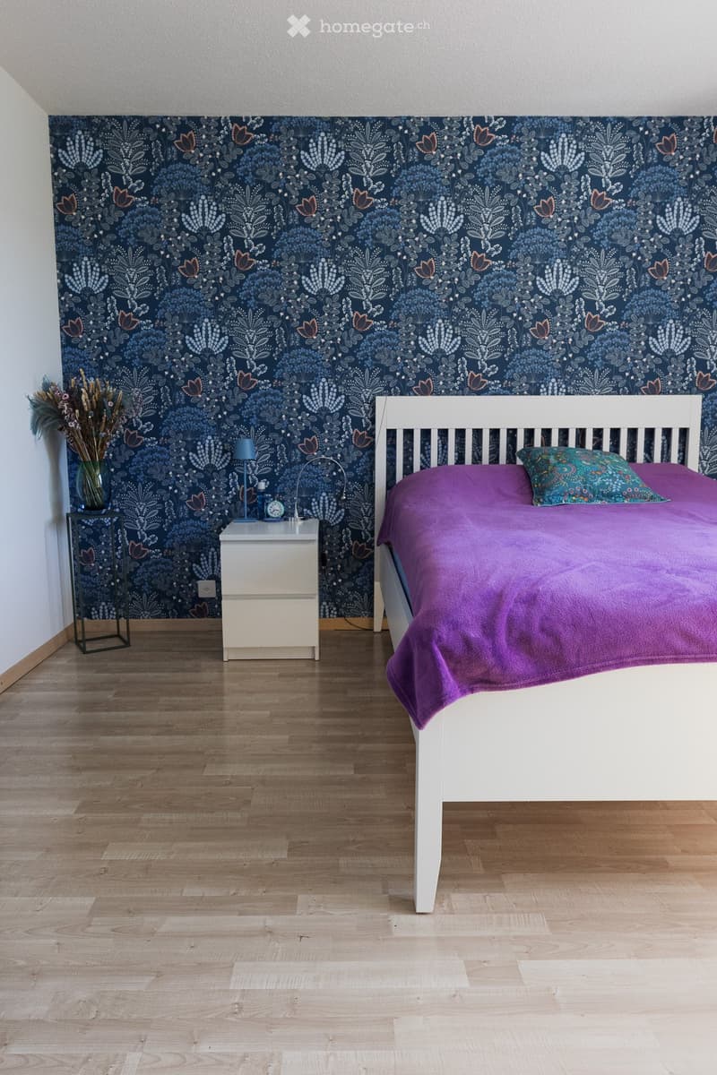 A modern bedroom with a white bed, purple bedding, a nightstand with a lamp, a vase with dried flowers, and a decorative wall mural.