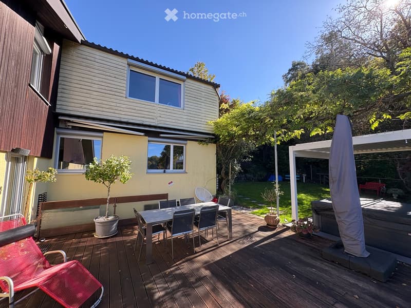 Wooden deck area with outdoor furniture, including a table, chairs, and a red lounge chair. The deck is adjacent to a house with yellow and white siding and large windows. There is a covered patio area with a grill and potted plants, surrounded by greener