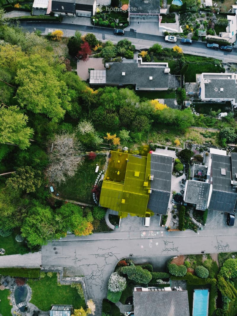 Aerial view of a residential neighborhood with multiple houses, gardens, driveways, and parked cars. The central house features a large, modern yellow-roofed structure with a swimming pool and landscaped garden.