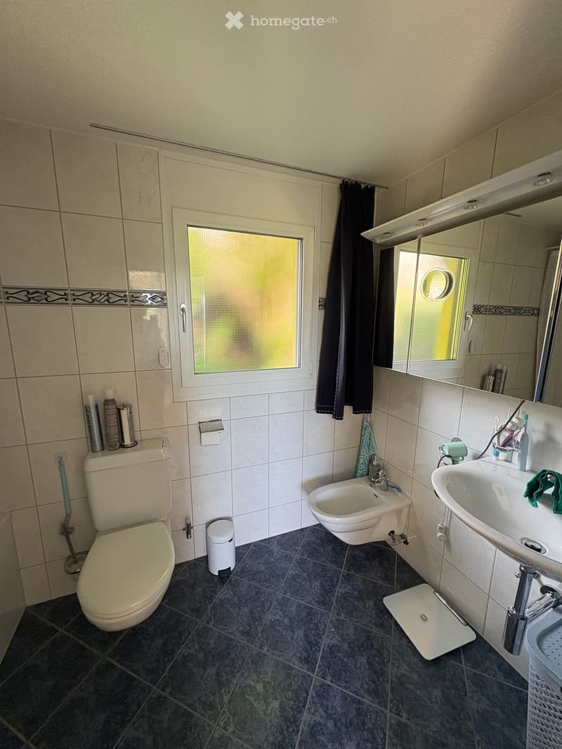 Bathroom with a toilet, sink, bidet, mirror, and a window with a curtain. The room features tiled walls and a dark tiled floor.