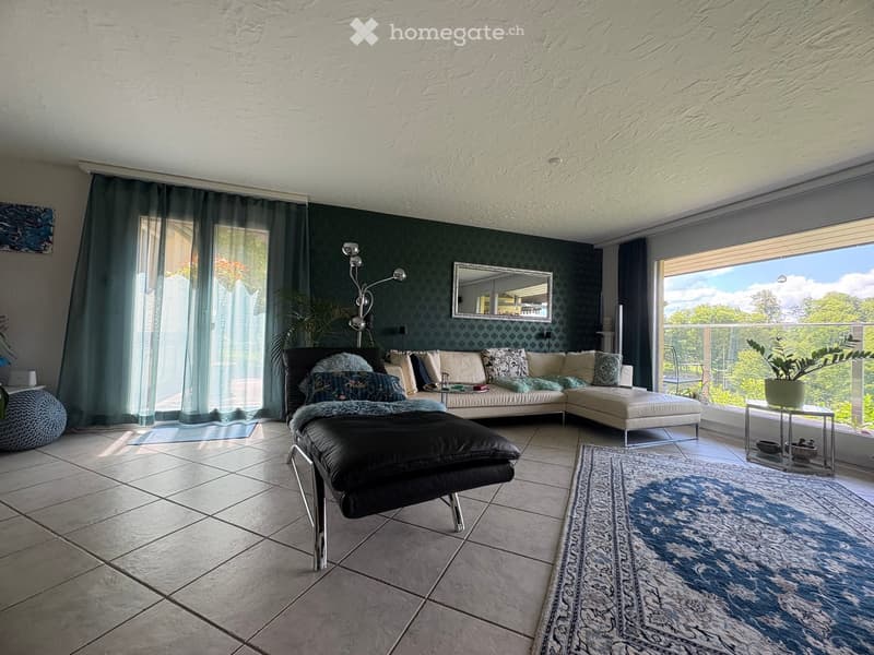 Modern living room with large windows offering a scenic view, featuring a cream-colored sectional sofa, black ottoman, coffee table, floor lamp, and decorative rug.