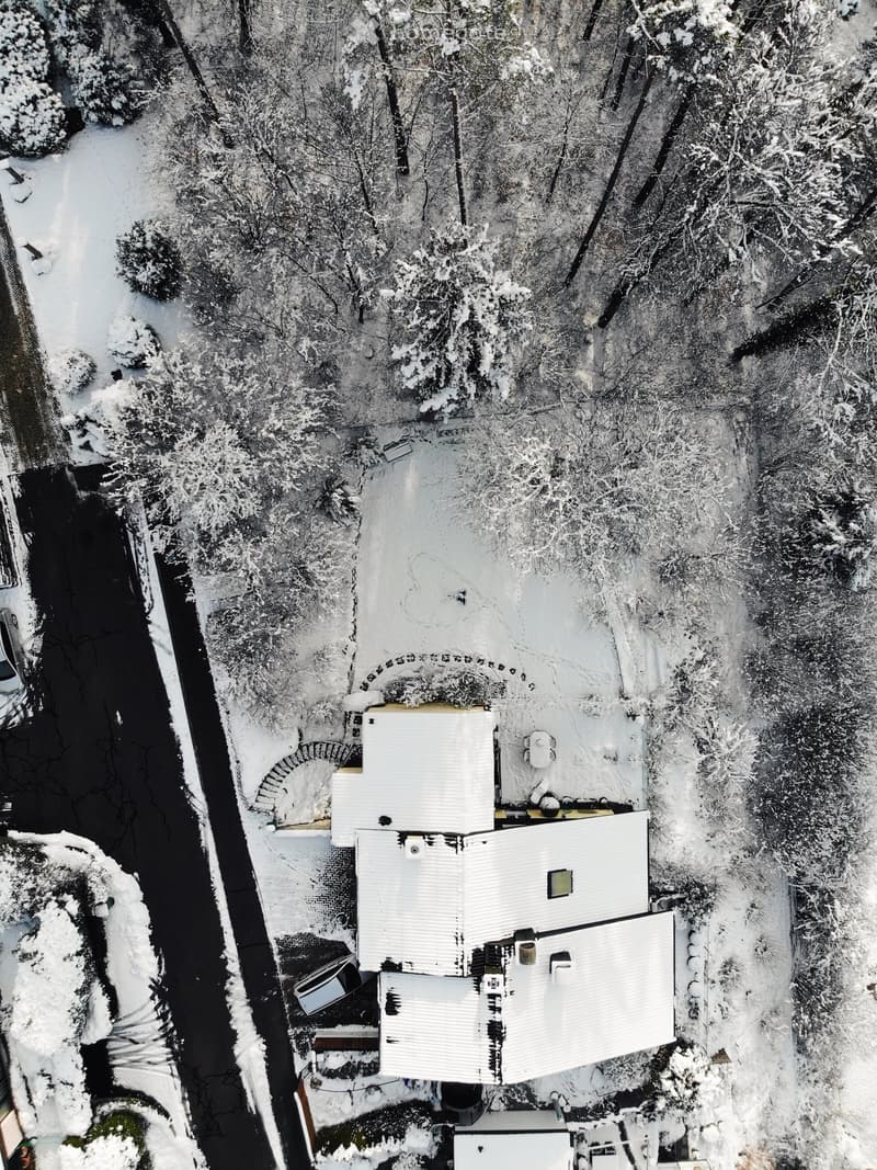 Aerial view of a house with a white roof and multiple sections, surrounded by snow-covered trees and a driveway with parked cars.