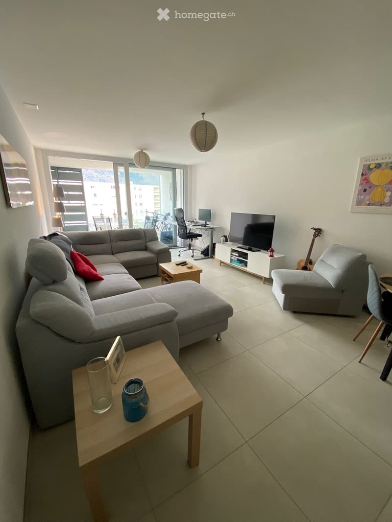 Modern living room with a grey sectional sofa, coffee tables, a guitar, a TV, and a view of the city through large windows.