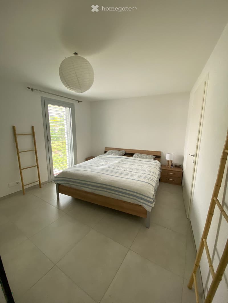 Bedroom with a wooden bed, striped bedding, a nightstand with a lamp, and a large window with blinds. The room has tiled flooring and white walls.