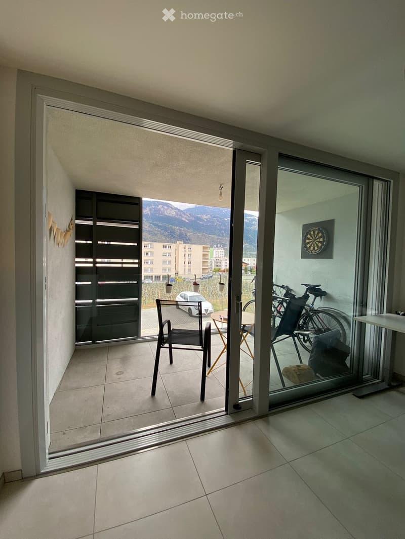 Balcony with tiled floor, sliding glass doors, outdoor furniture including a chair and small table, bicycles, and a view of mountains and buildings.