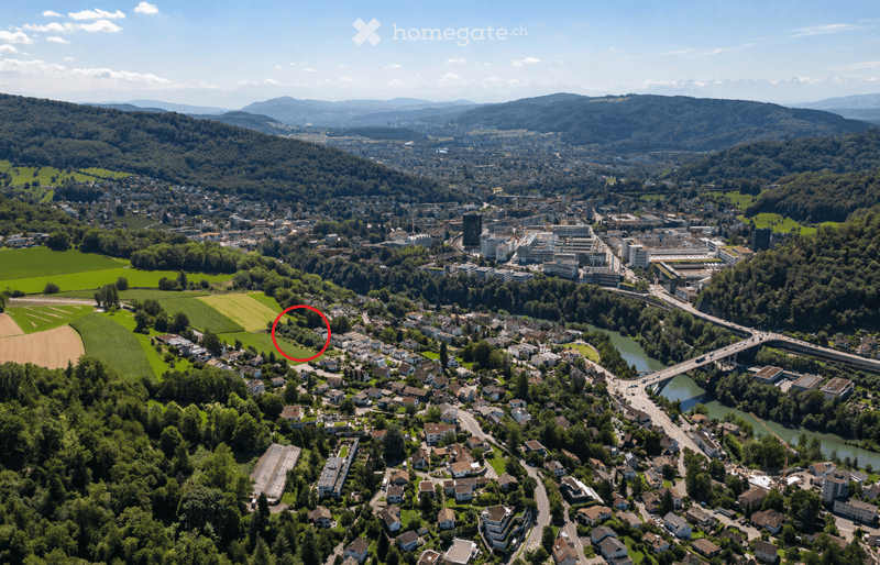 Aerial view of a town nestled in a valley surrounded by green hills and mountains, with a river running through it and a bridge crossing over it.