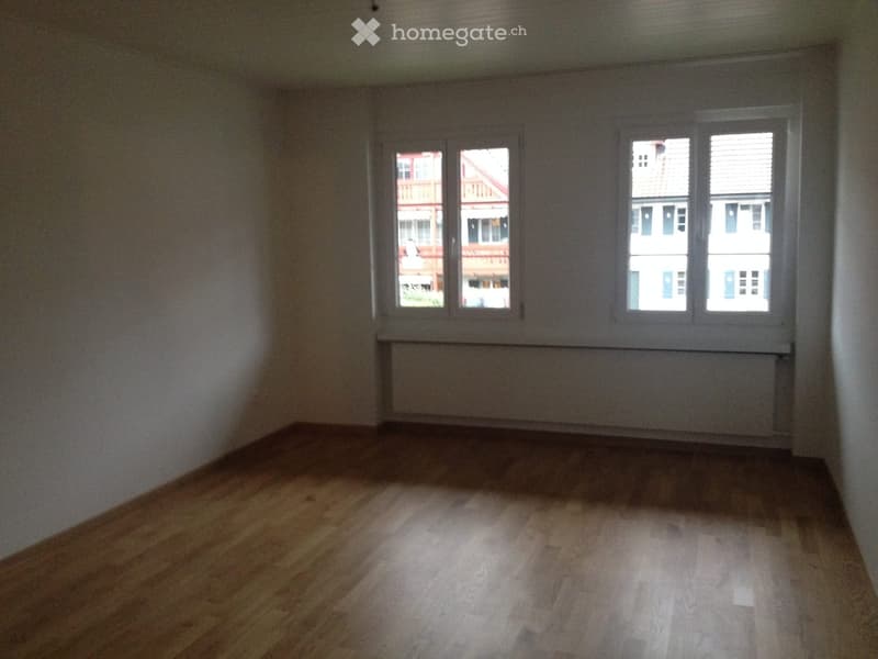 Empty room with wooden flooring, white walls, and large windows showing neighboring buildings.