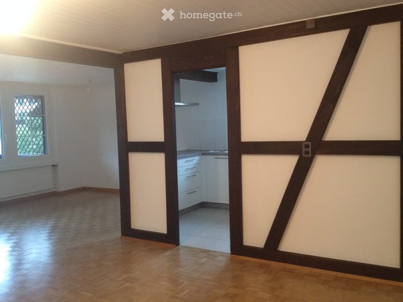 Empty room with wooden flooring, white walls, and a window with a metal security grid. A doorway leads to a small kitchen area with white cabinets and a countertop.