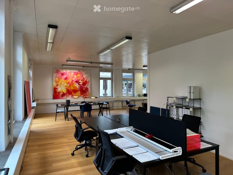 Office space with wooden flooring, multiple desks, chairs, a large screen, shelves, and artwork on the wall.