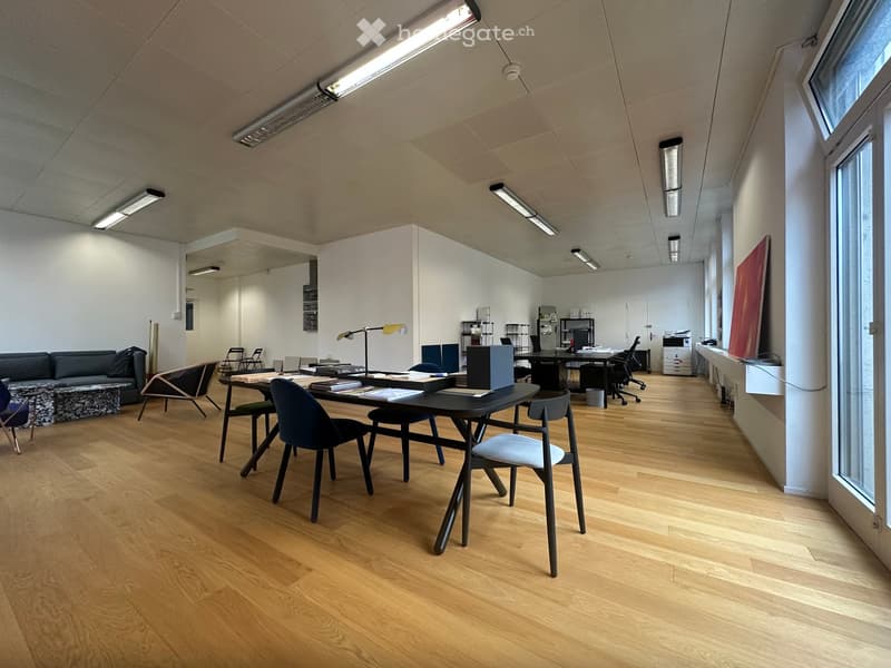 Modern office space with wooden flooring, large windows, and contemporary furniture including a sofa, desks, chairs, and a lamp.