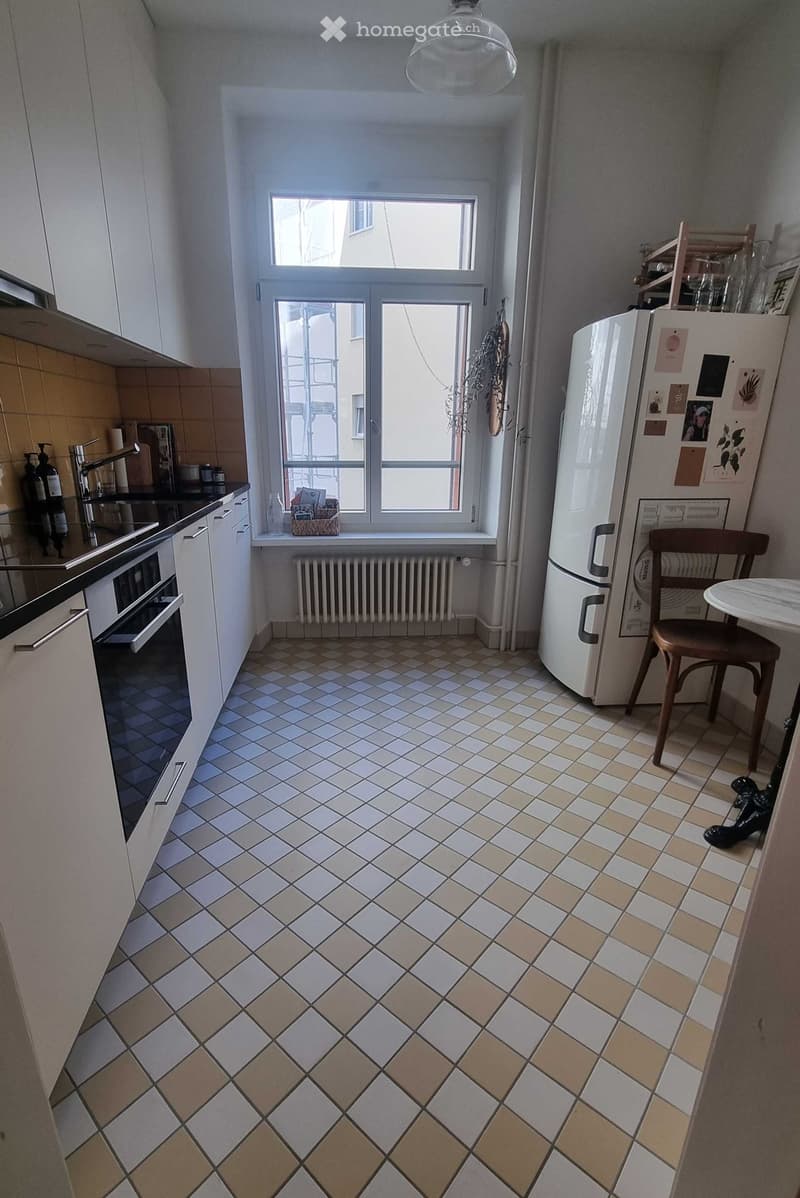 Modern kitchen with white cabinets, tiled floor, refrigerator, small table with chair, and window with view.