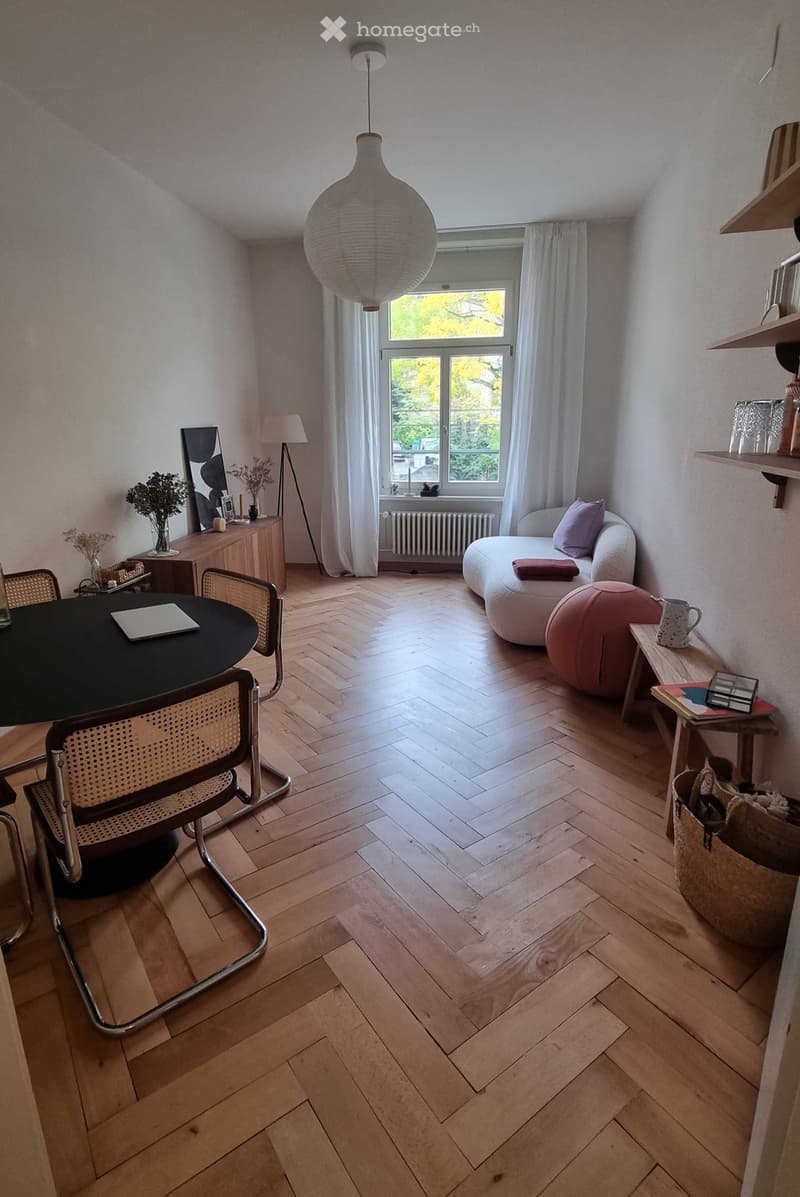 A modern living room with herringbone wooden flooring, a large window with white curtains, a hanging pendant light, a white sofa with a pink throw, a round black dining table with chairs, a small wooden shelf, and decorative items including vases and a pi