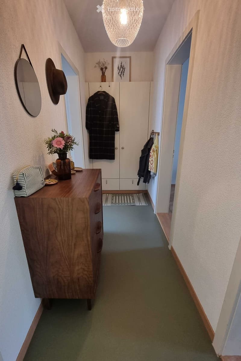 A narrow hallway with a wooden dresser on the left, a coat rack with coats and hats, and a hanging light fixture above. The walls are painted white, and the floor is carpeted in green.