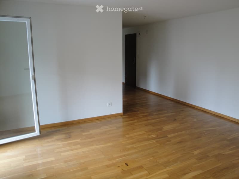 Empty room with light-colored walls and wooden flooring, featuring a large glass door and a small adjacent hallway.
