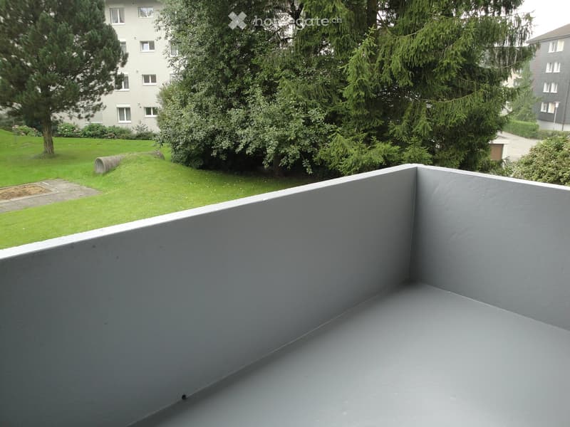 Grey concrete balcony with low walls, surrounded by greenery and apartment buildings.