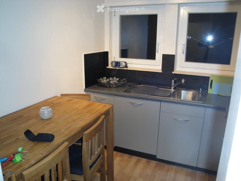 Modern kitchen with wooden dining table, chairs, stainless steel sink, grey cabinets, and a small radio on the counter.