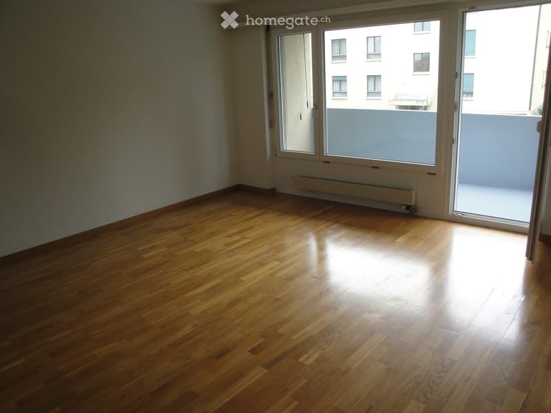 Empty room with wooden flooring, large sliding glass doors leading to a balcony, radiator on the wall, and a view of neighboring buildings.