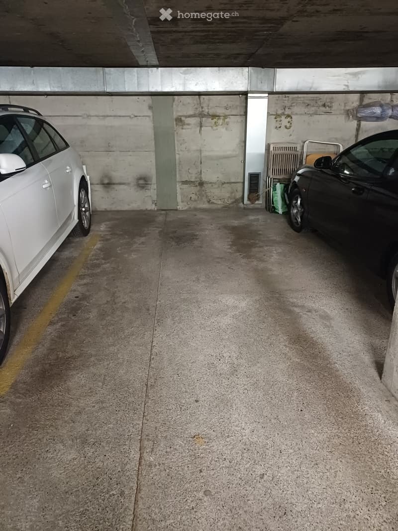 Underground parking area with two parked cars, a white car on the left and a black car on the right, concrete floor and walls, and a yellow parking line.