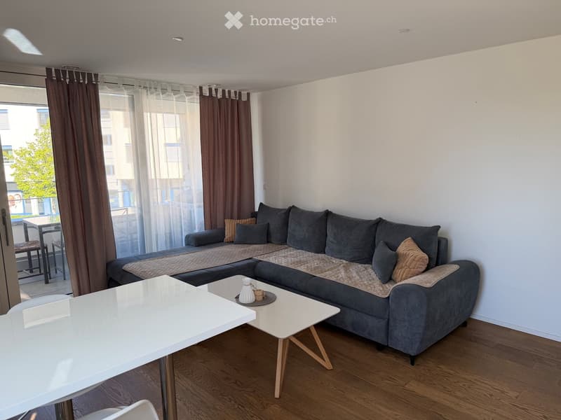 Modern living room with grey sectional sofa, two white coffee tables, and a white dining table. The room features wooden flooring, large windows with curtains, and access to a balcony.