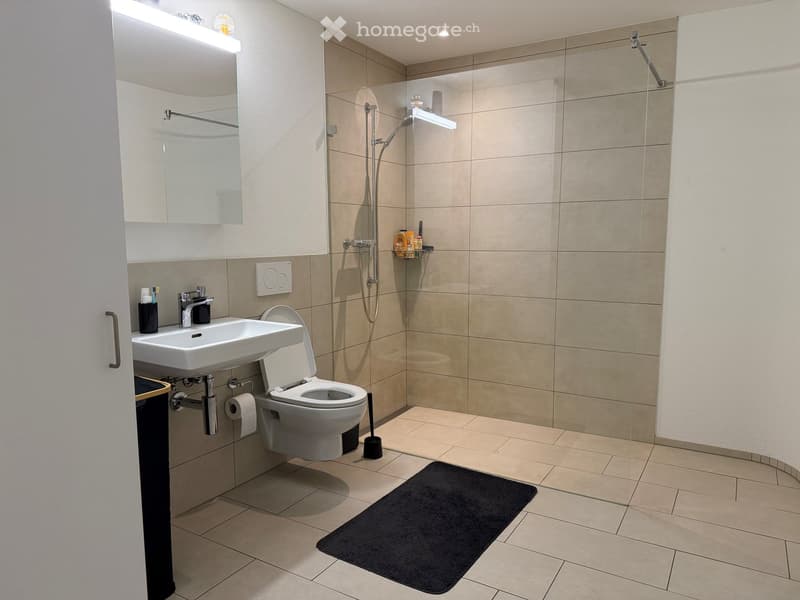 Modern bathroom with beige tiles, a white sink, toilet, shower with glass enclosure, and a black bathroom rug.