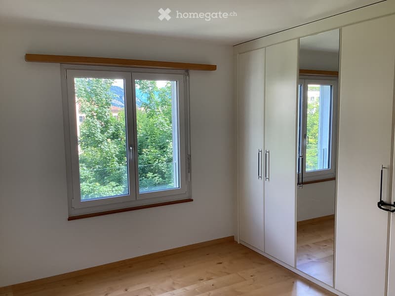 Empty room with light wooden flooring, white walls, large window with wooden sill, and built-in white wardrobe with mirrored doors.